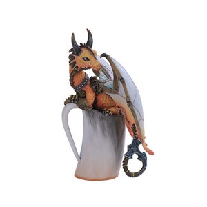May include: A small orange and brown dragon figurine perched on the rim of a white ceramic mug. The dragon has a long tail that wraps around the handle of the mug. The dragon's wings are spread out behind it.