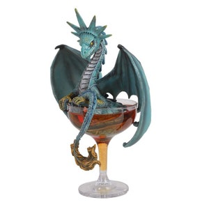 May include: A teal blue dragon figurine with gold accents sits inside a clear glass cocktail glass filled with a red liquid. The dragon has a spiky crest and wings.