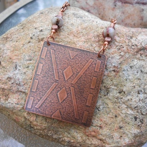 May include: A copper pendant necklace with a geometric design and white stone beads. The pendant is square shaped and has a textured surface.
