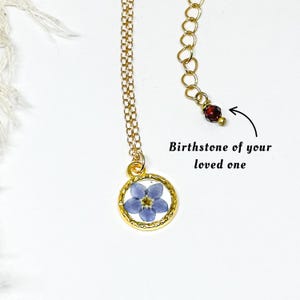May include: A gold-colored necklace with a small, round pendant containing a blue flower. The necklace has a delicate chain and a small red gemstone at the end. Text on the image reads "Birthstone of your loved one."