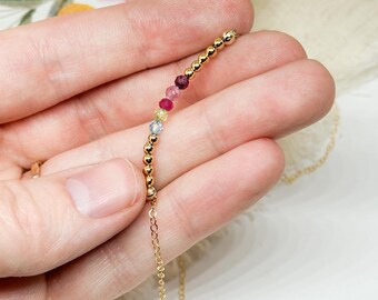 Puede incluir: Un delicado collar de oro con una hilera de pequeñas piedras preciosas de colores. El collar tiene una serie de cuentas doradas y una delicada cadena. Las piedras preciosas incluyen piedras rosas, rojas, amarillas y azules. El collar se sostiene en una mano.