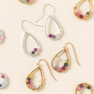 May include: A collection of teardrop-shaped earrings. Some are silver-toned, others gold-toned. Each earring features a wire-wrapped design holding a cluster of colorful gemstones, including pink, green, and blue stones. The earrings are displayed on a cream-colored surface.