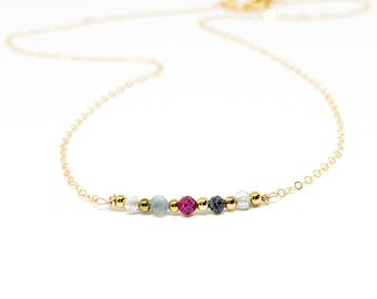 May include: A delicate gold chain necklace with a row of small, colorful gemstone beads. The beads are white, light blue, pink, dark blue, and white.