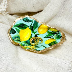 May include: A gold-rimmed ceramic dish with a lemon tree design. The dish is painted with green leaves and yellow lemons. Two gold rings are sitting in the dish.