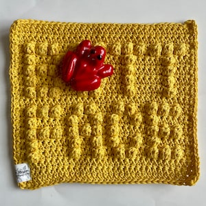 Puede incluir: Un paño de cocina de crochet amarillo con un patrón texturizado. Una figurita de rana roja está sentada encima del paño de cocina.