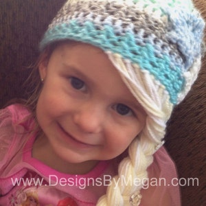 May include: A young girl wearing a blue, white, and gray crocheted hat with a long white braid attached to the side. The braid is styled to look like a princess's hair. The girl is wearing a pink shirt with a princess design.
