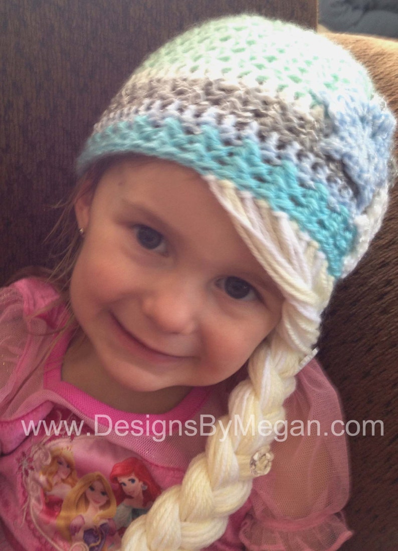 May include: A young girl wearing a blue, white, and gray crocheted hat with a long white braid attached to the side. The braid is styled to look like a princess's hair. The girl is wearing a pink shirt with a princess design.
