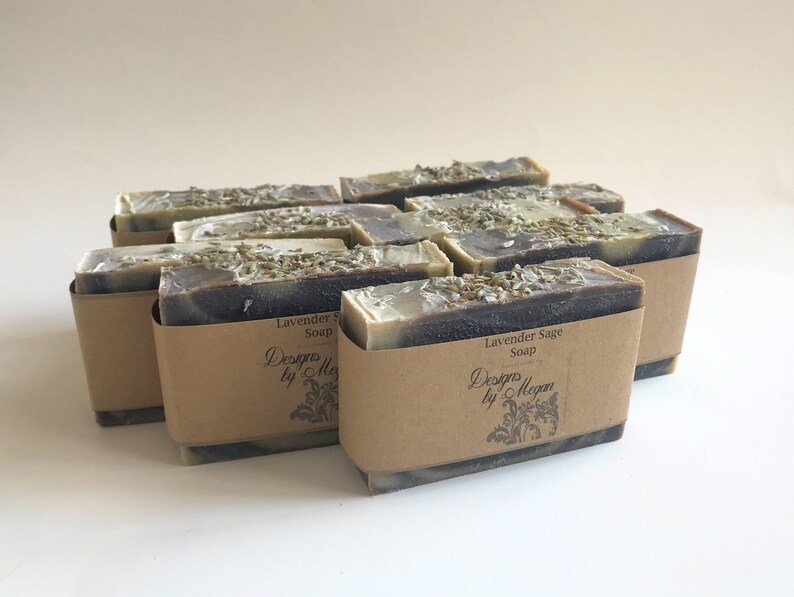 Pu&ograve; includere: Un mucchio di otto saponette alla lavanda e salvia avvolte in carta marrone con un'etichetta che recita "Designs by Megan" e "Lavender Sage Soap".
