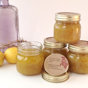 May include: A collection of small glass jars filled with yellow sugar scrub. The jars have gold-colored lids and are labeled. Lemons and a purple bottle are in the background. The label reads "Lavender Lemon Rosemary Sugar Scrub".