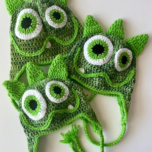 May include: Three green crocheted monster hats with large white-rimmed eyes and black pupils. The hats have pointed ears and dangling ties. The crochet work features varying shades of green, creating a textured appearance.