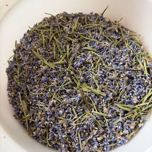 May include: A white bowl filled with a mixture of dried purple lavender buds and thin, light green dried rosemary needles. The contents are scattered evenly within the circular container, showing a contrast between the floral and herbal elements.
