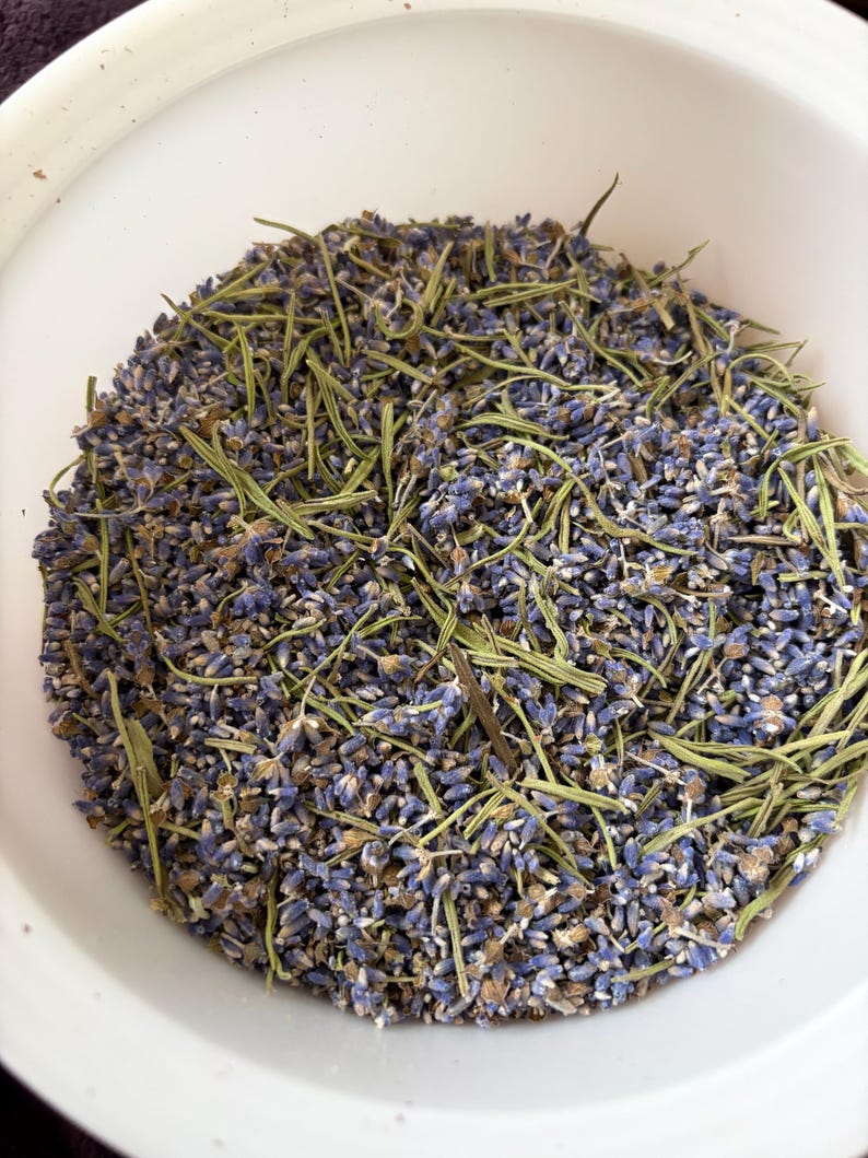 May include: A white bowl filled with a mixture of dried purple lavender buds and thin, light green dried rosemary needles. The contents are scattered evenly within the circular container, showing a contrast between the floral and herbal elements.