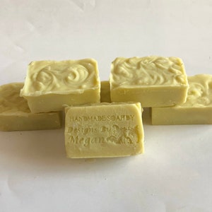 以下が含まれることがあります： 淡い緑色の手作り石鹸6個セット。各石鹸には、上面に装飾的な渦巻き模様があります。1つの石鹸には「HANDMADE SOAP BY Designs By Megan」の文字が刻印されています。