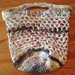 May include: A white crocheted bag made from recycled plastic bags. The bag has a handle and a round bottom. The bag is made with a variety of colors, including white, gray, brown, and red.