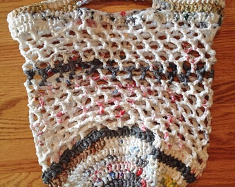 May include: A white crocheted bag made from recycled plastic bags. The bag has a handle and a round bottom. The bag is made with a variety of colors, including white, gray, brown, and red.