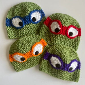 May include: Four green crocheted hats with different colored eye masks. The hats have white eyes with black pupils and are decorated with orange, red, blue, and purple eye masks.