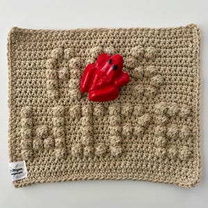 May include: A beige crocheted square with a textured bubble design and a vibrant red frog ornament. The frog has black eyes and is centrally placed. A small tag with text is in the lower left corner.