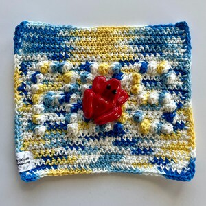 Puede incluir: Un paño de crochet cuadrado hecho a mano en tonos de azul, amarillo y blanco, con un patrón de puntadas en relieve texturizado. Una figura de rana roja brillante está centrada en el paño. El paño tiene una etiqueta en la esquina que dice "by Megan".