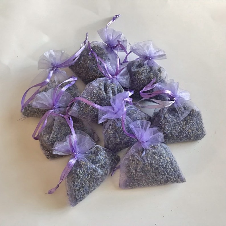 May include: A collection of small, semi-transparent light purple organza drawstring bags filled with dried lavender buds. Each pouch is cinched at the top with a matching lavender-colored satin ribbon, creating a gathered, ruffled effect.