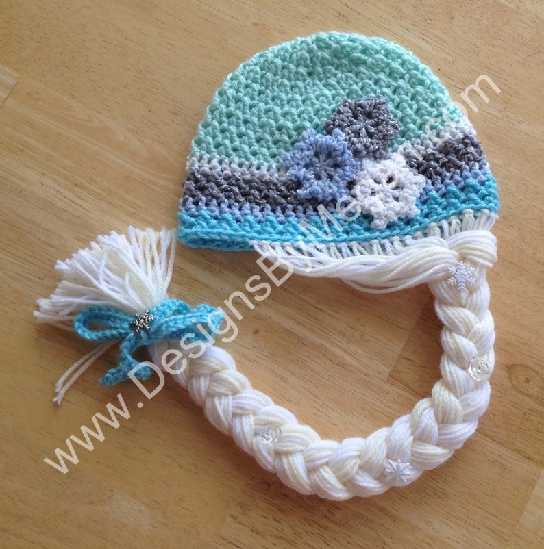 May include: A crocheted hat with a braid attached, the hat is blue, white, and gray with snowflake designs. The braid is white with white snowflake embellishments.