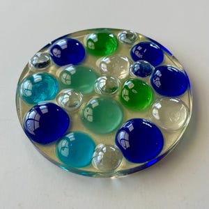 May include: A round, clear resin coaster with a collection of colorful glass beads. The beads are in shades of blue, green, and clear, creating a vibrant, textured surface. The coaster is a decorative item.