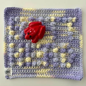 May include: A square, handmade crochet washcloth in shades of lavender, yellow, and gray, with a raised, textured pattern. A glossy red frog figurine is placed on the washcloth. The washcloth has a tag that reads "Megan megan.com".