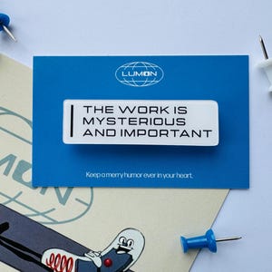 May include: A white rectangular pin with the text "THE WORK IS MYSTERIOUS AND IMPORTANT" on a blue card. The card has the Lumon logo and the text "Keep a merry humor ever in your heart." Blue and white push pins are scattered around.