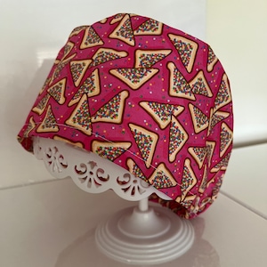 May include: Pink fabric headband with a pattern of yellow and brown triangles filled with colourful sprinkles. The headband is on a white display stand.