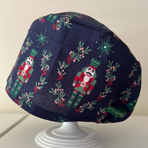 May include: A navy blue scrub cap with a festive Christmas pattern. The fabric features nutcrackers, sprigs of berries, and star-shaped ornaments in red, green, and white. A small green tag with the word "merry" is attached.