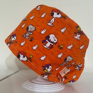 Może przedstawiać: Pomarańczowy czepek chirurgiczny na Halloween z Snoopy'm w różnych kostiumach, w tym mumii, wampira i pirata, wraz z białymi postaciami duchów. Czapka jest ręcznie robiona z metką z napisem "handmade with".