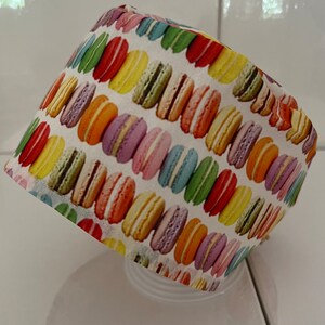 Op de afbeelding: Een witte scrubmuts van stof met een kleurrijk macaronpatroon. De muts is ontworpen voor gebruik in een medische omgeving.