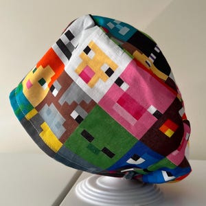 May include: A patterned scrub cap with pixelated character designs in orange, yellow, pink, green, brown, and blue. The fabric cap has a curved brim and is designed for medical or care settings.