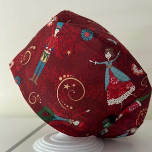 May include: A burgundy scrub cap featuring a repeating pattern of a toy soldier, a ballerina, and floral and swirl designs. The cap is made of fabric and is designed for medical use.