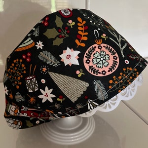 May include: A black scrub cap with a Christmas-themed pattern. The fabric displays white, pink, and orange floral and botanical designs. The cap has a white, scalloped edge.