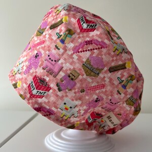 May include: Pink and white patterned cap featuring pixelated Minecraft characters, TNT blocks, and swords. The cap is made of fabric and has a small tag that says "Handmade".