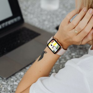 May include: A smartwatch with a pale pink strap and a gold-coloured case is worn on a person's wrist. The watch face shows a colourful image. A laptop is open in the background.
