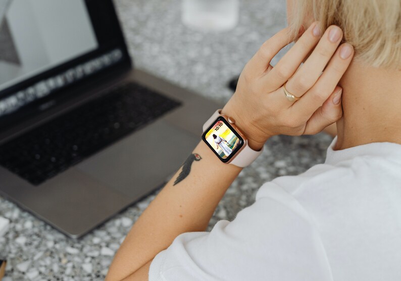 May include: A smartwatch with a pale pink strap and a gold-coloured case is worn on a person's wrist. The watch face shows a colourful image. A laptop is open in the background.