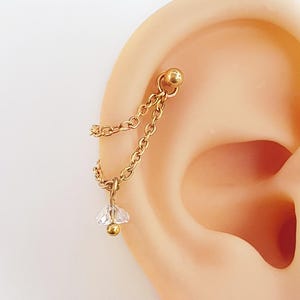 May include: Gold-coloured ear piercing jewellery featuring a chain and a small, clear, flower-shaped charm with a gold bead. The jewellery is attached to a gold ball stud. The earring is shown on a person's ear.