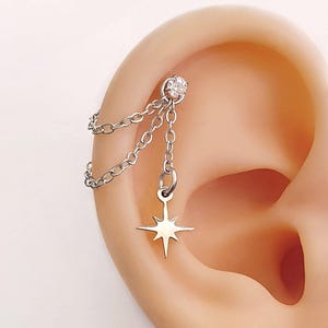 Peut inclure: Un piercing d'oreille argenté avec une chaîne et un charm en forme d'étoile. Le piercing a une petite gemme transparente en haut et est suspendu à une chaîne. Le charme en forme d'étoile a des bords pointus.