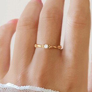 May include: A gold ring featuring a small, clear gemstone. The ring has an intricate, decorative design with a textured band. The ring is displayed on a finger, highlighting the jewellery's details and craftsmanship.