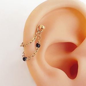 May include: A gold-colored ear piercing with a chain and black beads. The piercing is attached to the ear with a small ball stud. The chain dangles down from the stud, connecting to another bead. The jewelry is shown against a light skin tone.
