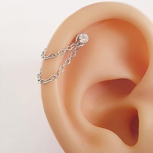 May include: A silver ear piercing with a small, clear gemstone stud linked by a fine chain to another chain. The jewellery is displayed on an ear, highlighting the piercing's design and features.