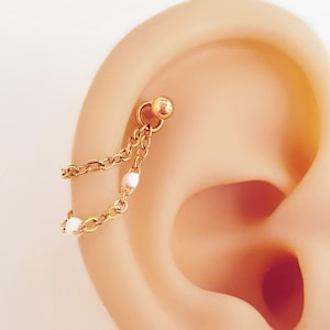 Könnte beinhalten: Goldfarbener Ohrschmuck mit Kettenmuster. Die Kette ist mit kleinen weißen Perlen verziert. Der Schmuck wird mit einem kleinen Kugelstecker am Ohr befestigt.