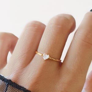 May include: A delicate gold ring with a heart-shaped gemstone. The ring has a textured band and a small, clear gemstone set in the centre. The ring is worn on a finger.