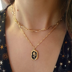 Puede incluir: Conjunto de collares dorados. La cadena superior tiene eslabones ovalados, mientras que la cadena inferior tiene un colgante con fondo negro y diseño floral dorado. El colgante es de forma ovalada.