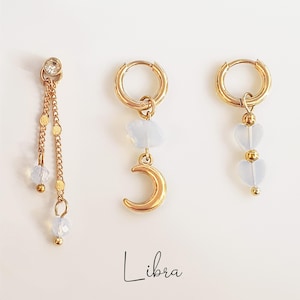 May include: Gold-coloured earrings with celestial and heart-shaped charms. One earring has a dangling chain with beads, while the others have a moon and heart design. The word "Libra" is written in cursive.