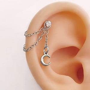 Può includere: Un piercing all'orecchio argentato con un piccolo borchia di pietra preziosa trasparente e un ciondolo a forma di mezzaluna pendente. Il ciondolo è collegato alla borchia con delicate catene, creando un effetto a strati. Il gioiello è mostrato su un orecchio.
