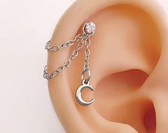 May include: A silver-toned ear piercing featuring a small, clear gemstone stud and a dangling crescent moon charm. The charm is connected to the stud with delicate chain links, creating a layered effect. The jewelry is shown on an ear.