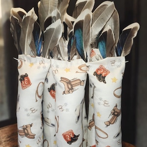 May include: A collection of decorative feather arrangements. Each arrangement features a bundle of gray and blue-green feathers emerging from a white fabric pouch. The pouches are printed with a Western-themed pattern of boots, saddles, and stars.