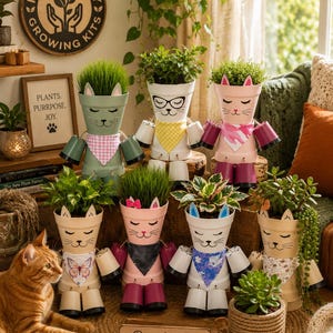 May include: Seven cat-shaped planters made from stacked terracotta pots, each featuring painted faces, ears, and fabric bandanas. The planters hold various green plants and are arranged in two rows. A small wooden sign reads Rooted in love. Growing with joy.